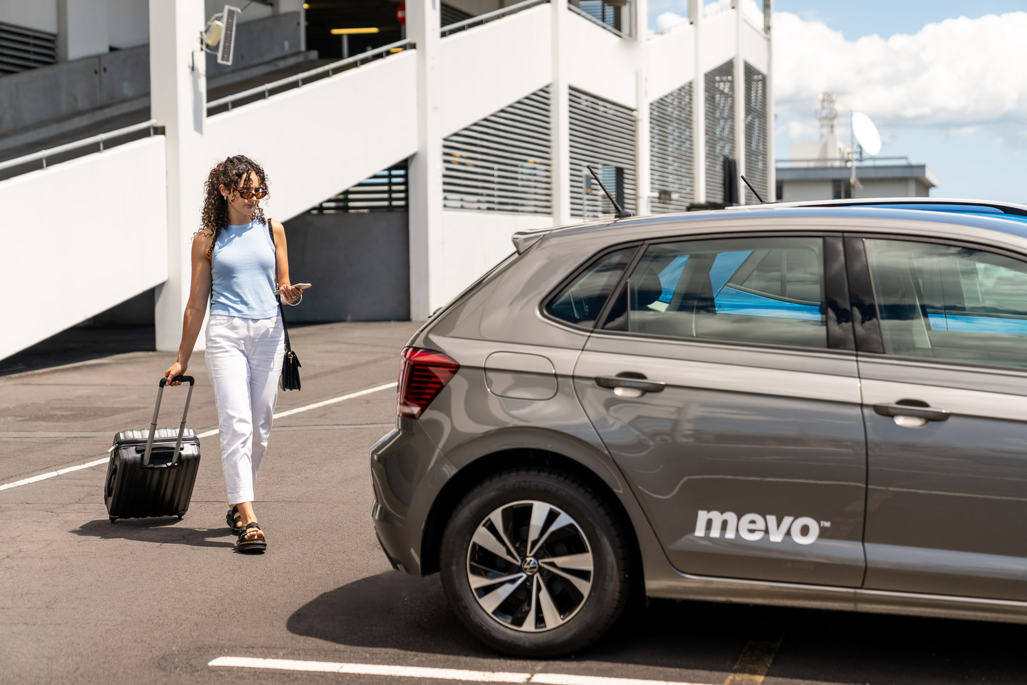 Woman at Auckland Airport walking towards a Mevo car, pulling a suitcase