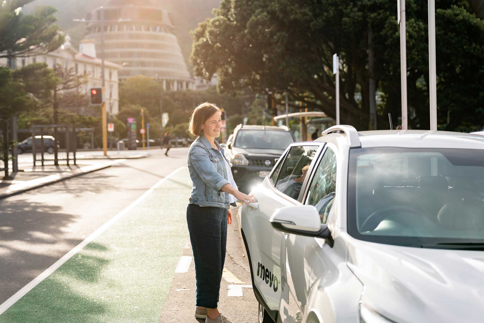 Ursula opening the rear door of a Mevo vehicle on a Wellington street; the Beehive is visible in the background.